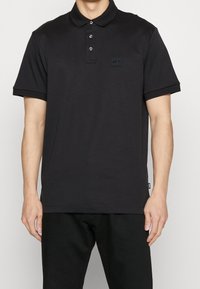 Svart pikétröja med krage, tre knappar i knäppningen och en liten logotyp på vänster sida. Korta ärmar med snygg finish.