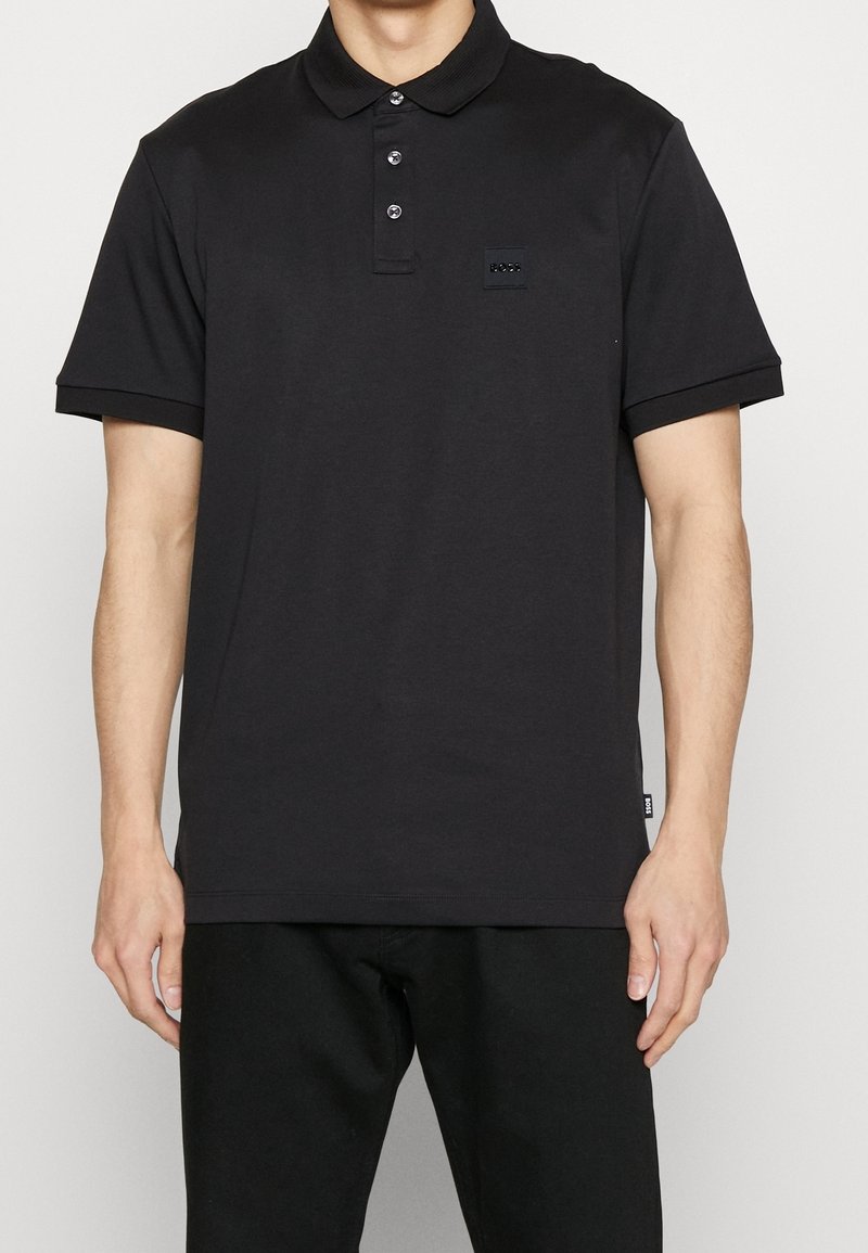 Svart pikétröja med krage, tre knappar i knäppningen och en liten logotyp på vänster sida. Korta ärmar med snygg finish.