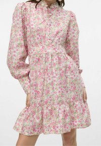 Kvinna i en rosa blommig klänning med lång puffärmar, en vågig nederkant och en hög knäppning vid halsen, stående med händerna på höfterna.