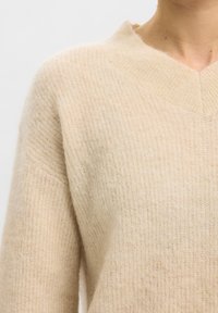 Pull en maille douce beige avec un large col en V côtelé porté par une personne visible de l'épaule jusqu'au milieu de la poitrine.