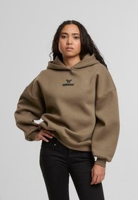 Khaki színű, oversized kapucnis pulóver, textúrált felülettel, fekete logóval a mellkason és nagy kapucnival. Slim fit fekete farmernadrággal párosítva.