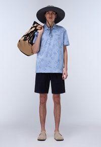 Ljusblå polo med lövmönster, mörka shorts, bredskärmad grå hatt och beige slip-ons, som håller en brun och svart väska.