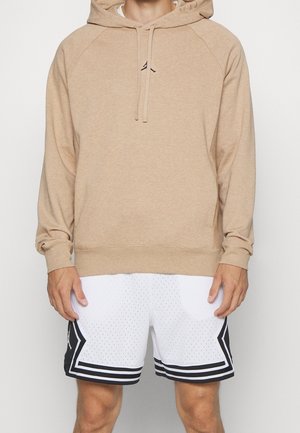 Mand iført en beige hættetrøje og hvide basketballshorts med sorte striber, stående med hænderne afslappet ned langs siderne.