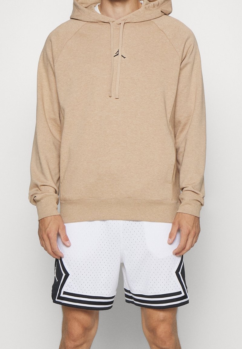 Homme portant un sweat à capuche beige et un short de basket blanc avec des rayures noires, debout les mains détendues le long du corps.
