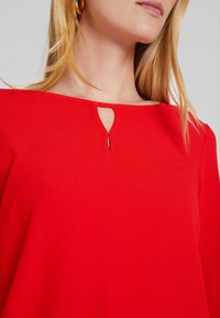 Blouse rouge texturée avec un décolleté bateau et un petit détail en forme de clé. Tissu lisse, manches longues et coupe décontractée.