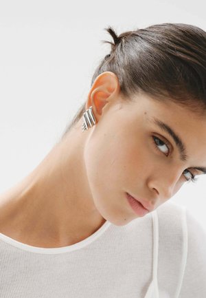 Pendientes de plata con un diseño ondulado y bandas de metal en capas, exhibidos en un modelo sobre un fondo neutro.