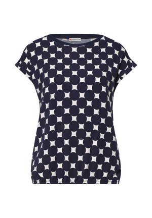 Kvinde iført kortærmet navy og hvid polkaprikket skjorte med opkrogede ærmer og lyseblå jeans, vist bagfra, smilende let.