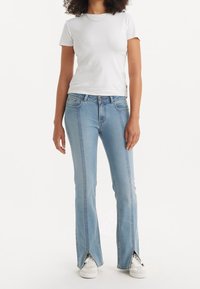 Jeans de mezclilla azul claro con un diseño acampanado, que presentan aberturas laterales en el dobladillo y costuras en contraste. Combinados con una camiseta blanca lisa y zapatillas deportivas.