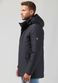 Tumši zils izolēts jaka ar kapuci, ar rāvējslēdzēja kabatu, teksturētu audumu un pieguļošu formu. Logo uzlīme uz kreisā piedurknes.