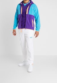 Giacca in fleece colorata con pannelli blu e viola, dettagli arancioni e tasche con zip; abbinata a pantaloni jogger bianchi e scarpe da ginnastica bianche.