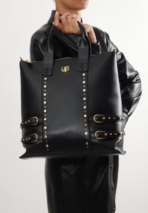 Borsa tote in finta pelle nera con dettagli in metallo dorato, caratterizzata da accenti di borchie e dettagli con fibbie sui lati; include doppi manici superiori e chiusura con cerniera.