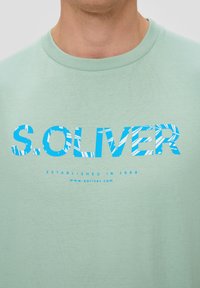 Hellgrünes Baumwoll-T-Shirt mit fett blauen Buchstaben "S. OLIVER" in einem gestreiften Muster. Enthält "ESTABLISHED IN 1969" unter dem Logo.