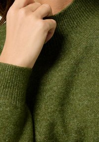 Zielony sweter z dzianiny z wysokim, prążkowanym kołnierzem i miękką fakturą, na zbliżeniu ręka dotykająca dekoltu.
