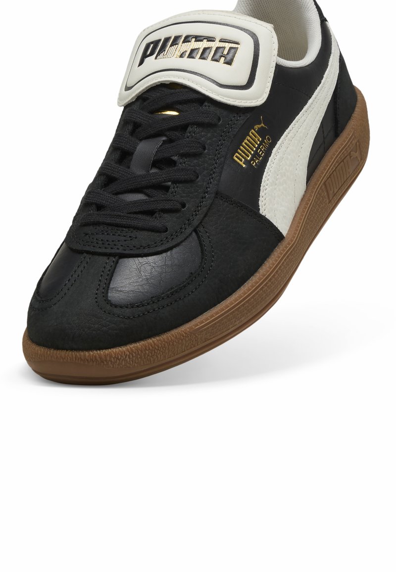 Baskets en cuir noir avec une bande blanche, semelle en caoutchouc, et languette rembourrée proéminente affichant le logo Puma en or. Tige texturée.