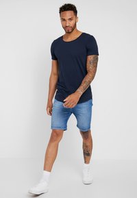 Maglietta a maniche corte blu scuro abbinata a pantaloni shorts di jeans chiari. Modella che indossa sneakers bianche e calzini alla caviglia. Tatuaggi visibili sulle braccia della modella.