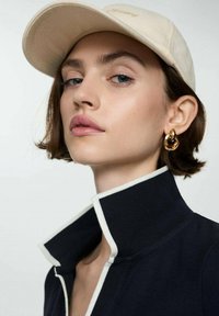 Beige kasket med logo, marineblå jakke med hvid kant, guld hoopsørenge og et nærbillede af en kvindes ansigt med neutral makeup og lys hud.