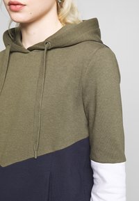Sweatshirt com capuz apresentando blocos de cor em verde-azeitona e azul-marinho, textura suave, cordões ajustáveis e mangas brancas em contraste.