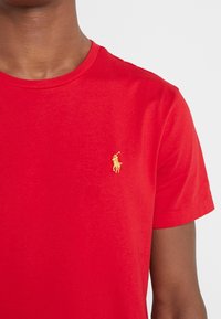 Rood katoenen T-shirt met een ronde hals en korte mouwen; heeft een klein geel geborduurd paardenlogo op het linkerborstgebied.