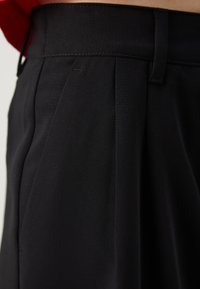 Black trousers featuring a flat waistband, pleated front, and smooth fabric texture. The design emphasizes structured tailoring and versatility.
