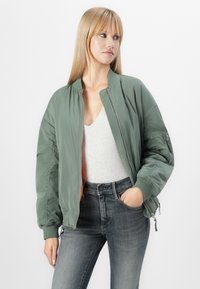 Giubbotto bomber verde oliva con zip, polsini a coste e interno arancione. Abbinato a una maglietta grigia a scollo a V e jeans grigio scuro.