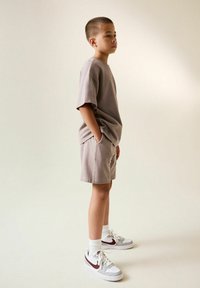 Béžové bavlnené tričko s krátkymi rukávmi, doplnené o zodpovedajúce šortky. Biely a šedý tenisky s bordovými akcentmi a rebrované biele ponožky.