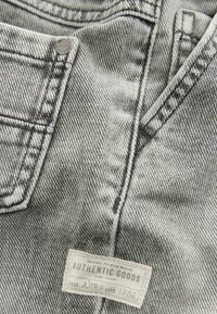 Jean gris avec un tissu en denim texturé, un design à cinq poches et une étiquette portant la mention "AUTHENTIC GOODS" près de la taille.