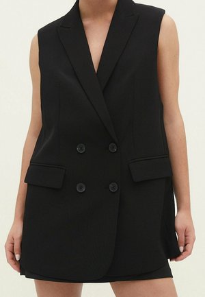 Blazer noir sans manches à double boutonnage avec revers crantés et poches à rabat porté sur une jupe ou un short noir.
