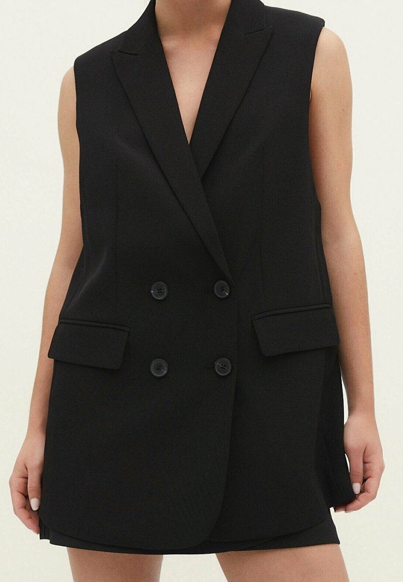 Blazer noir sans manches à double boutonnage avec revers crantés et poches à rabat porté sur une jupe ou un short noir.