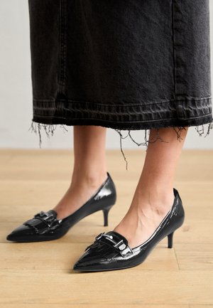 Kvinde fødder iført skinnende sorte spidse kitten heels med frynser og spænde detaljer, stående på lyst trægulv.