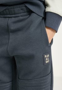 Tmavě modré teplákové kalhoty z měkké látky s texturovaným povrchem, prošitými detaily a čtvercovou nášivkou s nápisem "PLAY ALL DAY".