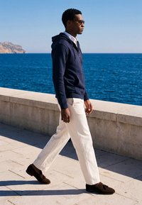 Homme portant des lunettes de soleil, un sweat à capuche bleu marine, un pantalon blanc et des chaussures marron marchant le long d'une promenade en bord de mer avec l'océan bleu et un ciel clair en arrière-plan.