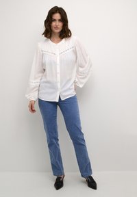 White button-up blouse with a V-neck, puff sleeves, and lace detailing across the chest. Worn with blue straight-leg jeans and black shoes.