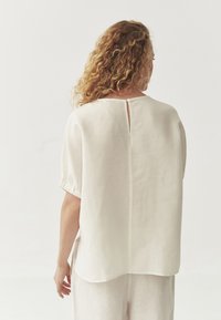 White linen top with short, puffed sleeves and a keyhole detail at the back. Flowy design, smooth texture, and slightly curved hem.
