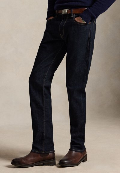 Muž nosi tmavomodré džínsy, hnedý kožený opasok, tmavý sveter a hnedé kožené topánky, stojí so založenými rukami v vreckách na jednoliatom pozadí.