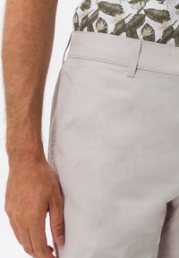 Pánská ruka odpočívající vedle světle béžových kalhot se boční kapsou, oblečena v bílé košili s potiskem zelených tropických listů.