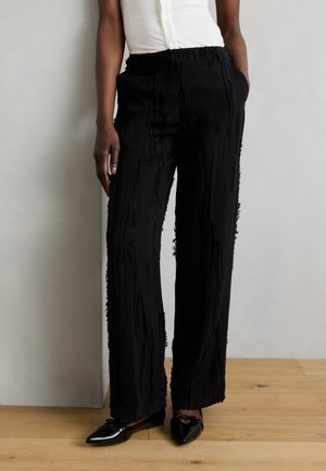 Persona che indossa pantaloni neri a gamba larga con texture, top bianco senza maniche e ballerine nere a punta, in piedi su pavimento in legno contro una parete bianca.