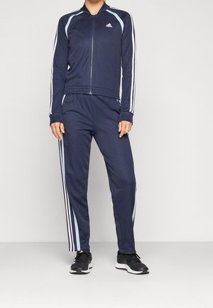 Osoba ubrana w granatowy dres Adidas z białymi paskami na rękawach i nogawkach, stojąca na jednolitym tle.