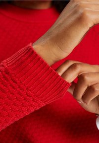 Pull rouge avec un corps texturé et des poignets côtelés. Gros plan sur une main ajustant le poignet, mettant en valeur les détails du tissu et l'ajustement.