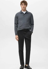 Młody mężczyzna stojący, ubranego w szarą bluzę polo z długim rękawem, czarne spodnie i czarne mokasyny, z jedną ręką w kieszeni.