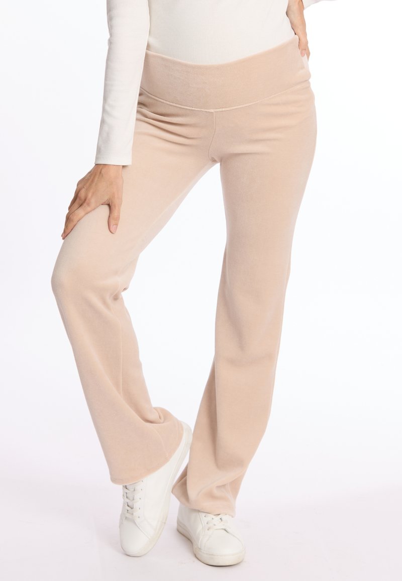 Pantalons évasés beiges avec une texture douce et veloutée, dotés d'une large taille. Associés à des baskets blanches, sur un fond blanc uni.