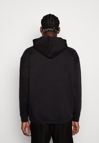 Felpa nera con cappuccio capiente, realizzata in materiale morbido. Presenta una vestibilità comoda e polsini a costine. La vista posteriore rivela un design minimalista.