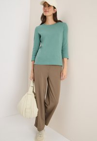 Haut long à manches longues teal clair associé à un pantalon large marron ; portant un sac blanc doux et un béret beige, avec des baskets.