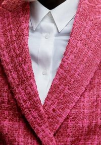 Vaaleanpunainen teksturoitu neuletakki, jossa on leveä kaulus, on kerroksittain valkoisen napitettavan paidan päällä, jossa on klassinen kaulus ja nappisulku.