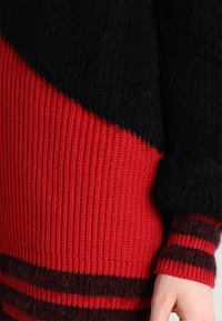 Stickad tröja med en svart och röd färgblockdesign, med ribbad struktur och randiga detaljer på ärmarna. Varmt och texturerat tyg.