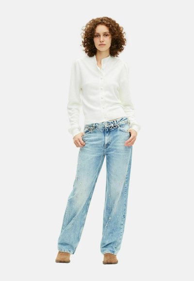 Femme aux cheveux bouclés portant un cardigan blanc à boutons, un jean large bleu clair avec des strass, une ceinture décorative en argent et des chaussures marron, debout.