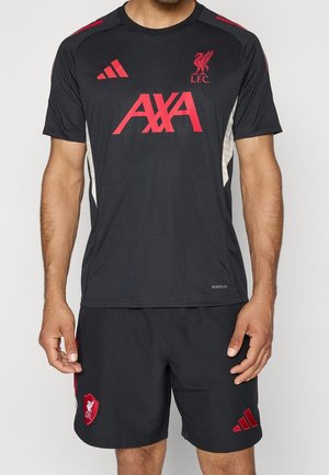 Mężczyzna w czarnym stroju treningowym Liverpool FC z czerwonymi logo Adidas i AXA, stoi na jednolitym jasnym tle.