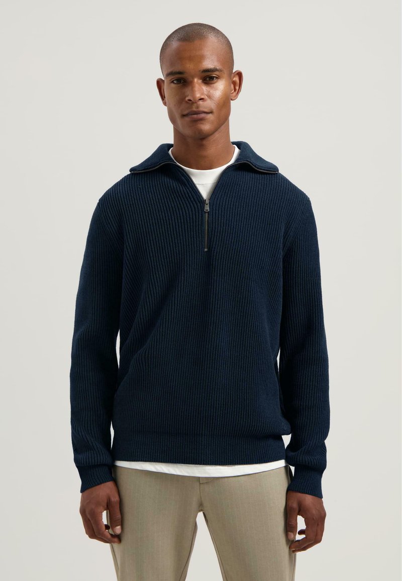 Man in een marineblauwe ribgebreide half-zip trui over een wit shirt en beige broek, staand tegen een effen lichte achtergrond.