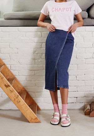 Vrouw in een roze "CHAPOT" t-shirt, denim midi rok met een vooropening, roze sokken en witte sandalen, staande bij houten trappen en een witte stenen muur.