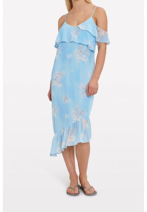 Woman wearing a light blue floral cold-shoulder dress with ruffles and gray flip-flops, standing against a plain background.