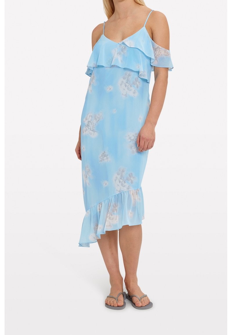 Femme portant une robe bleu clair fleurie à épaules dénudées avec des volants et des tongs grises, debout devant un fond uni.